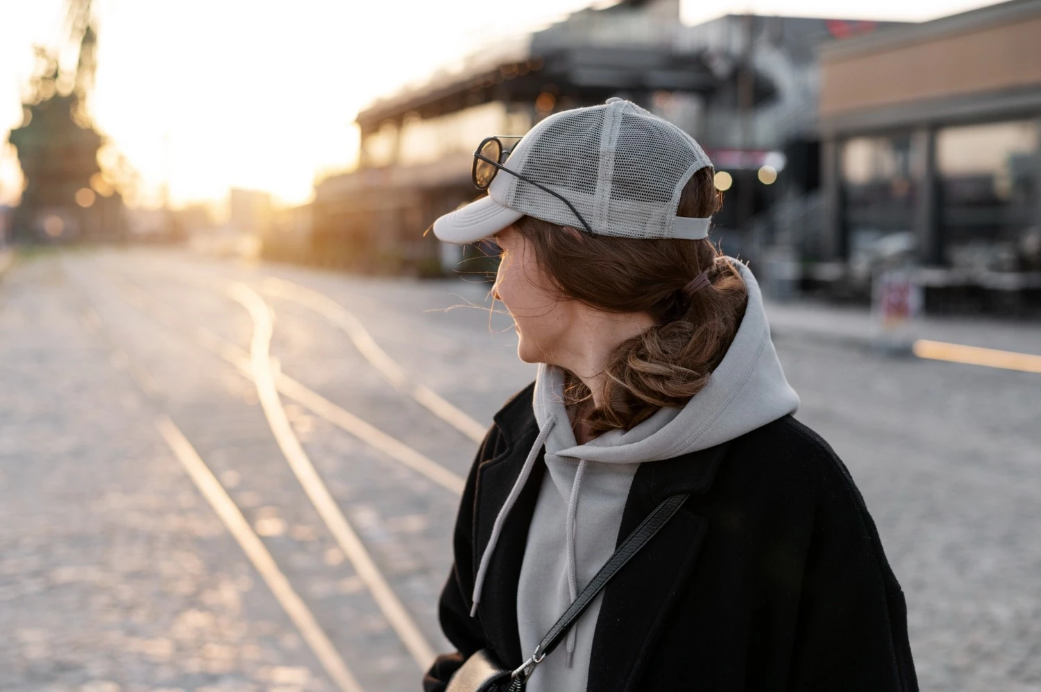 夕暮れ時の街路に立つ、灰色のトラッカーハットとサングラスをかぶった若い女性の横顔。