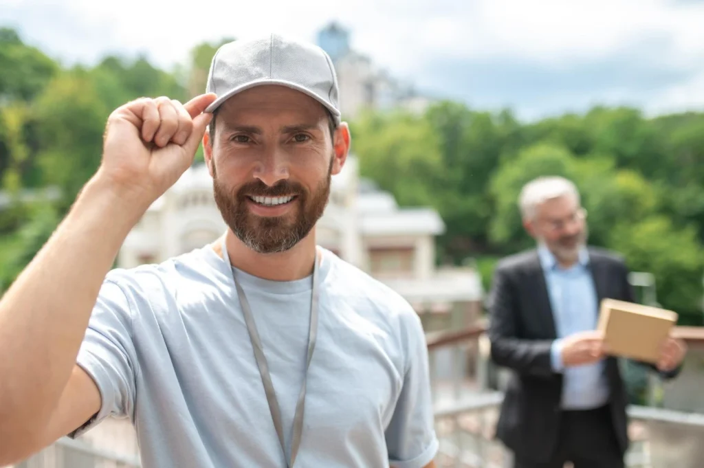 Man wearing a grey baseball cap and smiling outdoors with another person blurred in the background holding a box