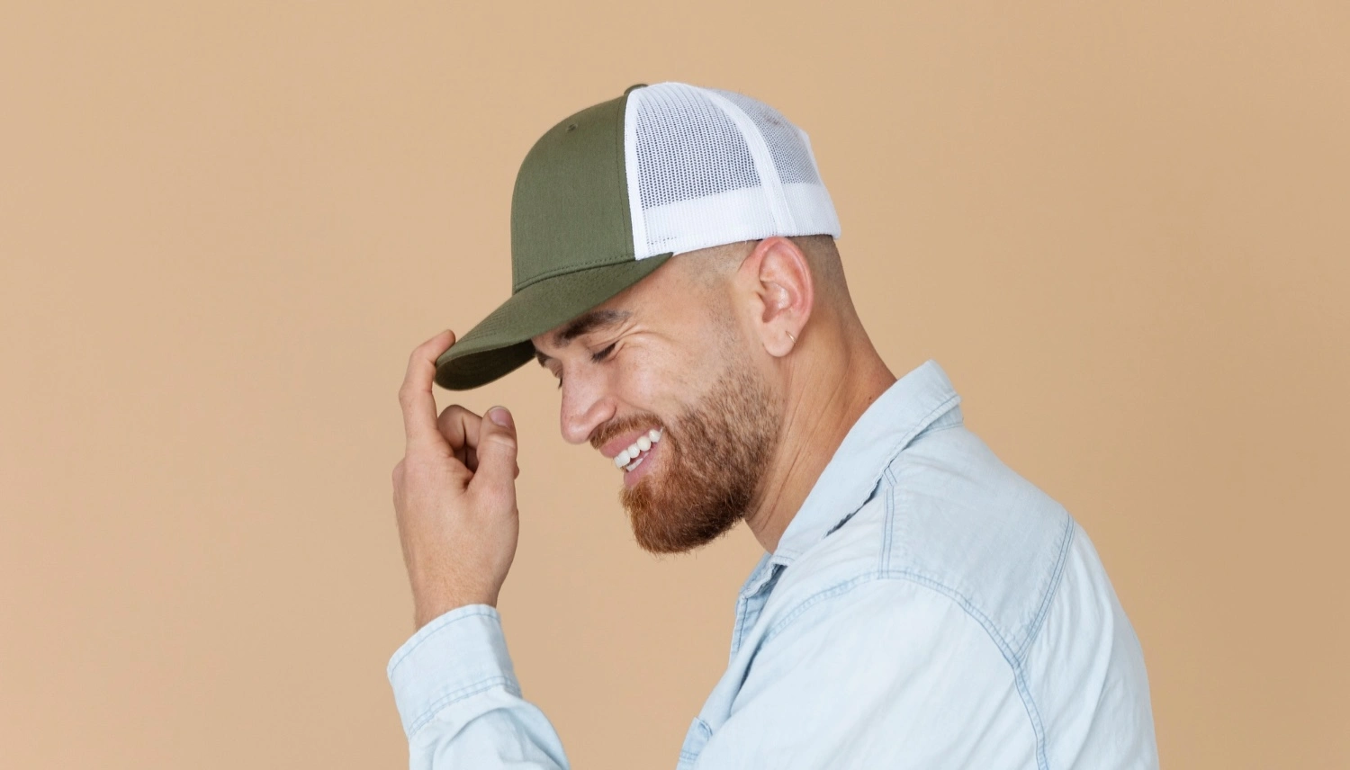 Smiling bearded man adjusting his green and white trucker cap against a beige background Smiling bearded man adjusting his green and white trucker cap against a beige background