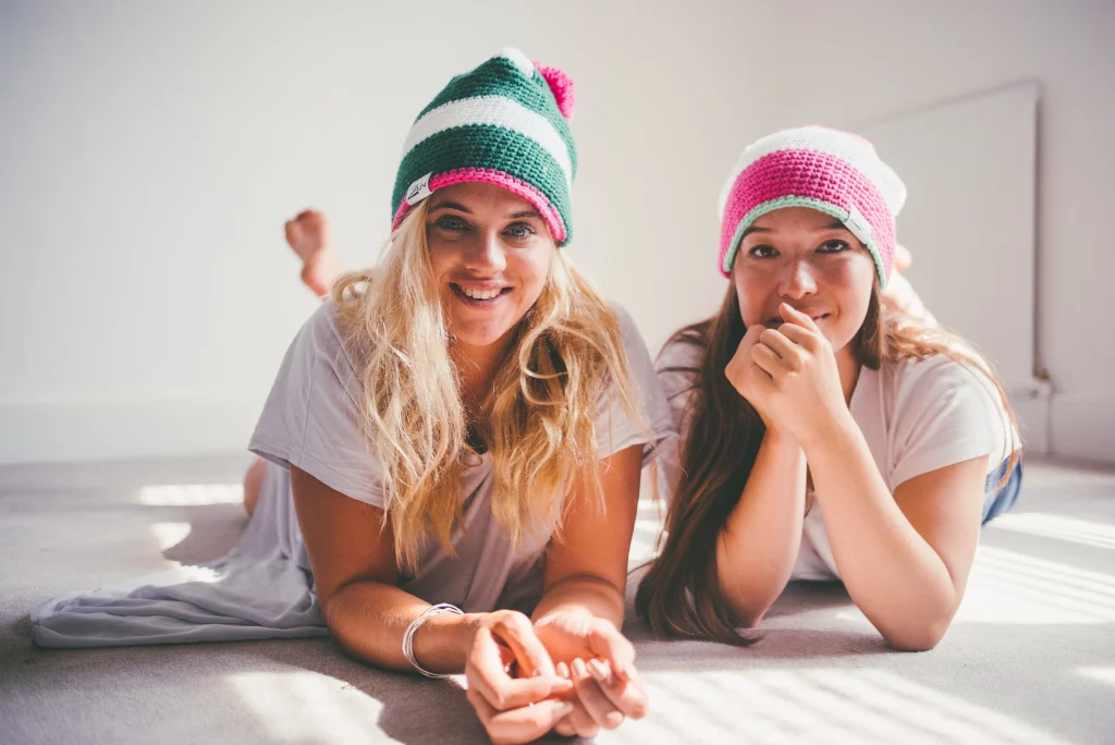 Two women in colorful crochet beanies smiling indoors
