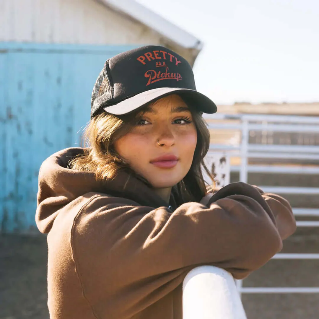 Woman in Pretty as a Pickup trucker hat leaning on a fence