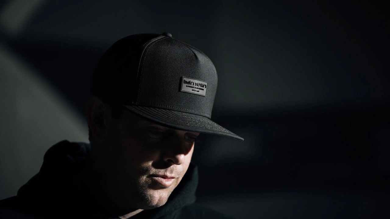 A man in a shadowy setting dons a classic black melin hat