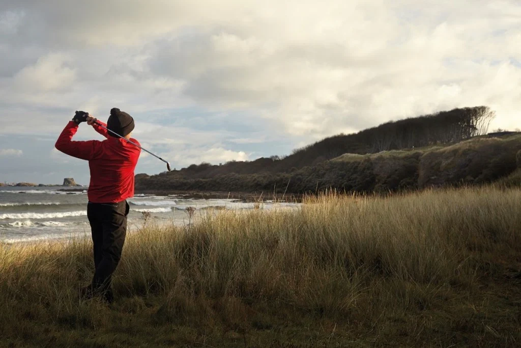 A golfer wearing a beanie swings near the coastline A golfer wearing a beanie swings near the coastline